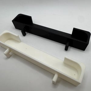 May include: Two rectangular, 3D-printed objects. One is black, the other is white. Both have a trough-like design with raised ends and a central support. The objects are likely components or parts for a larger assembly.