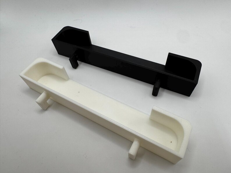 May include: Two rectangular, 3D-printed objects. One is black, the other is white. Both have a trough-like design with raised ends and a central support. The objects are likely components or parts for a larger assembly.