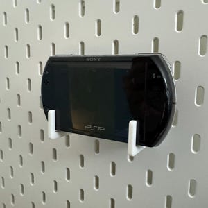 May include: A black Sony PSP handheld game console mounted on a white pegboard using white brackets. The PSP is labeled with the Sony logo and the letters "PSP". The pegboard has a pattern of evenly spaced, vertical slots.