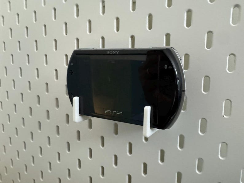 May include: A black Sony PSP handheld game console mounted on a white pegboard using white brackets. The PSP is labeled with the Sony logo and the letters "PSP". The pegboard has a pattern of evenly spaced, vertical slots.