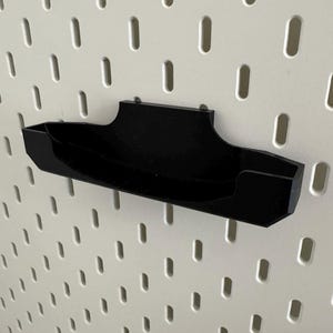 Op de afbeelding: Een zwarte, wandgemonteerde organizer van plastic met een gebogen ontwerp. De organizer is gemonteerd op een wit prikbord.