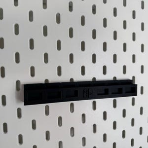 K&ouml;nnte beinhalten: Schwarzes Kunststoffregal zur Aufnahme einer Spielekonsole, montiert an einer wei&szlig;en Lochwand. Das Regal hat eine rechteckige Form mit mehreren F&auml;chern und dem Konsolenlogo.