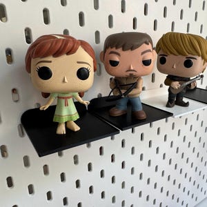 Pode incluir: Tr&ecirc;s figuras de vinil pop expostas em prateleiras pretas montadas em uma parede de placa de pinos branca. As figuras s&atilde;o uma menina com cabelo ruivo usando um vestido verde, um homem com cabelo castanho usando uma camisa preta e jeans azul, e um homem com cabelo loiro usando uma camisa marrom e jeans azul.