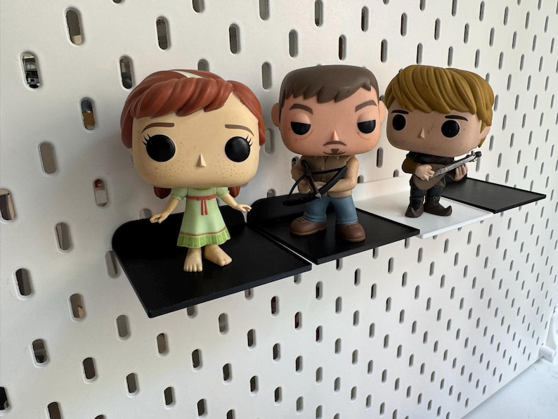 Pode incluir: Tr&ecirc;s figuras de vinil pop expostas em prateleiras pretas montadas em uma parede de placa de pinos branca. As figuras s&atilde;o uma menina com cabelo ruivo usando um vestido verde, um homem com cabelo castanho usando uma camisa preta e jeans azul, e um homem com cabelo loiro usando uma camisa marrom e jeans azul.