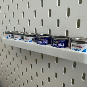 May include: A white shelf holds six small paint cans against a white pegboard. The cans are silver with blue and white labels, some labeled "Revell Email Color" and "Humbrol Enamel". Each can contains 14 ml or 0.47 fl oz of paint.