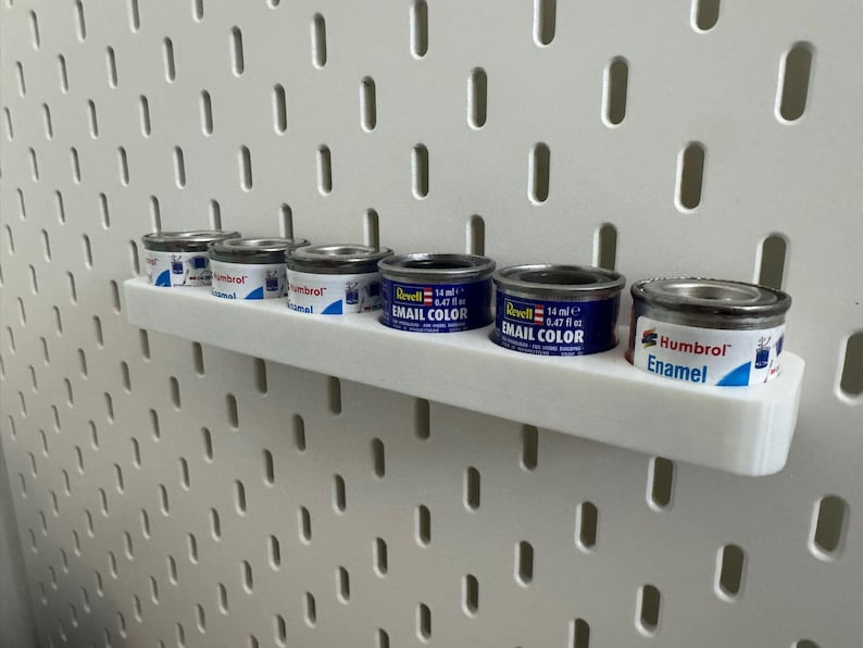 May include: A white shelf holds six small paint cans against a white pegboard. The cans are silver with blue and white labels, some labeled "Revell Email Color" and "Humbrol Enamel". Each can contains 14 ml or 0.47 fl oz of paint.
