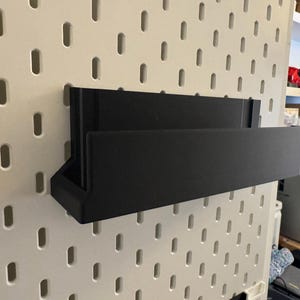 Peut inclure: Une &eacute;tag&egrave;re de rangement murale noire de forme rectangulaire. L'&eacute;tag&egrave;re est faite d'un mat&eacute;riau solide et est fix&eacute;e sur un panneau perfor&eacute; blanc avec des fentes espac&eacute;es r&eacute;guli&egrave;rement. L'&eacute;tag&egrave;re a un petit compartiment inclin&eacute; sur le c&ocirc;t&eacute; gauche.