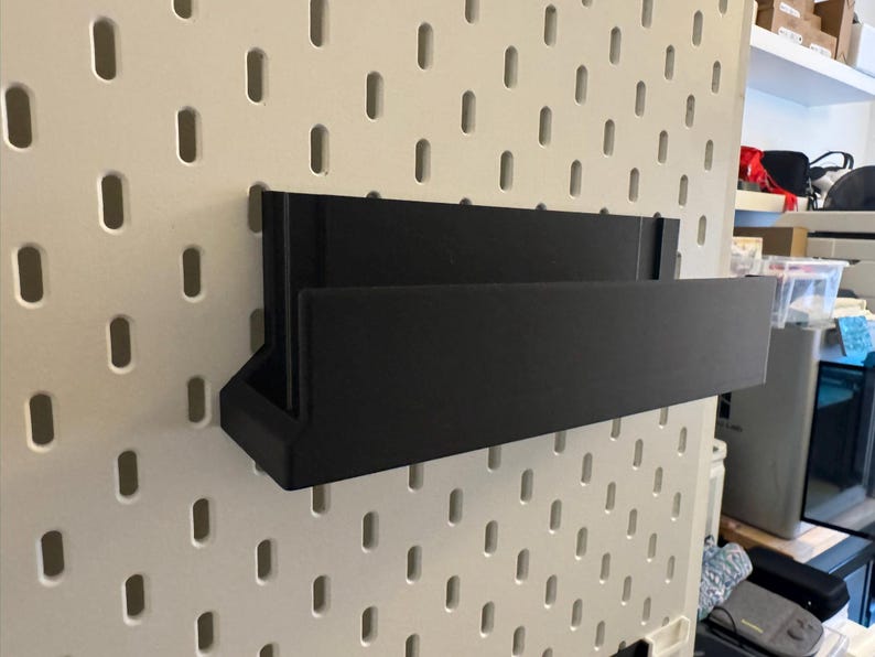 Peut inclure: Une &eacute;tag&egrave;re de rangement murale noire de forme rectangulaire. L'&eacute;tag&egrave;re est faite d'un mat&eacute;riau solide et est fix&eacute;e sur un panneau perfor&eacute; blanc avec des fentes espac&eacute;es r&eacute;guli&egrave;rement. L'&eacute;tag&egrave;re a un petit compartiment inclin&eacute; sur le c&ocirc;t&eacute; gauche.