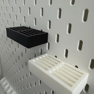 Op de afbeelding: Zwarte en witte rechthoekige pegboardplanken. De planken hebben een sleufontwerp en zijn gemonteerd op een wit pegboard. De zwarte plank bevindt zich boven de witte plank. Het pegboard heeft gelijkmatig verdeelde ovale gaten.