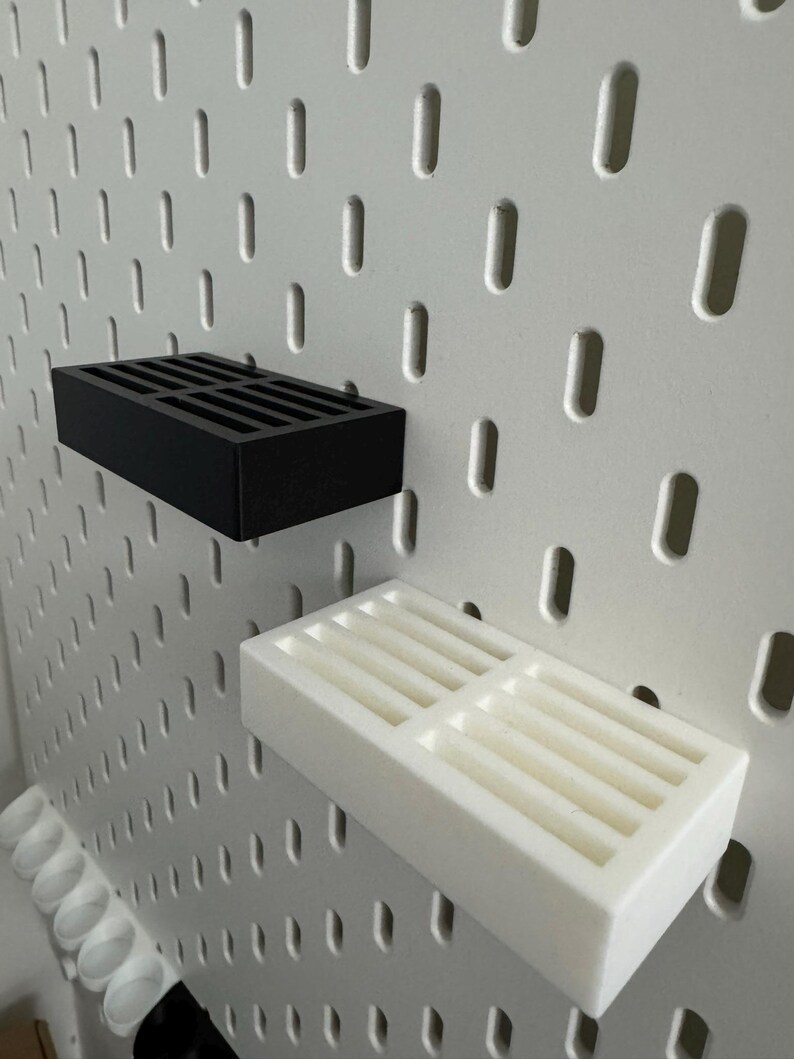 Op de afbeelding: Zwarte en witte rechthoekige pegboardplanken. De planken hebben een sleufontwerp en zijn gemonteerd op een wit pegboard. De zwarte plank bevindt zich boven de witte plank. Het pegboard heeft gelijkmatig verdeelde ovale gaten.