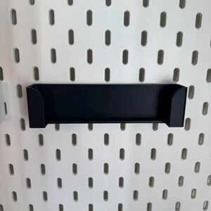May include: A black, rectangular shelf with curved sides, designed to attach to a white pegboard. The shelf is made of a solid material and is mounted on a white pegboard with evenly spaced oval holes.