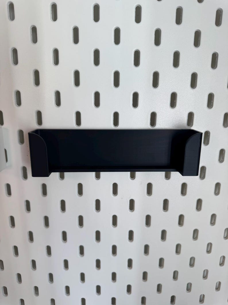 May include: A black, rectangular shelf with curved sides, designed to attach to a white pegboard. The shelf is made of a solid material and is mounted on a white pegboard with evenly spaced oval holes.