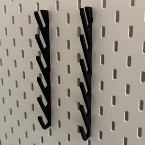 Pu&ograve; includere: Due ganci neri per pannello forato con pi&ugrave; punte. I ganci sono montati su un pannello forato bianco.