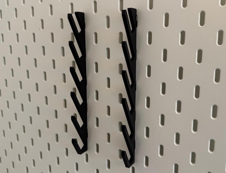 Pu&ograve; includere: Due ganci neri per pannello forato con pi&ugrave; punte. I ganci sono montati su un pannello forato bianco.