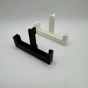 Puede incluir: Dos soportes para tel&eacute;fono impresos en 3D. Uno es negro, el otro es blanco. Cada soporte tiene una base rectangular con un soporte vertical y dos soportes laterales. Los soportes est&aacute;n dise&ntilde;ados para sostener un tel&eacute;fono en posici&oacute;n vertical.