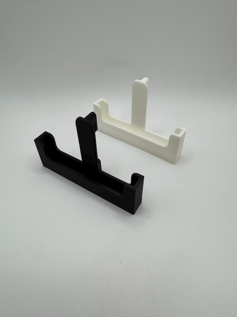 Puede incluir: Dos soportes para tel&eacute;fono impresos en 3D. Uno es negro, el otro es blanco. Cada soporte tiene una base rectangular con un soporte vertical y dos soportes laterales. Los soportes est&aacute;n dise&ntilde;ados para sostener un tel&eacute;fono en posici&oacute;n vertical.