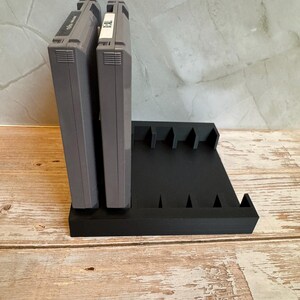 May include: Black, 3D-printed video game cartridge holder. The holder has slots for multiple cartridges and is designed to hold them upright. Two gray game cartridges are in the holder. The holder is on a wooden surface.
