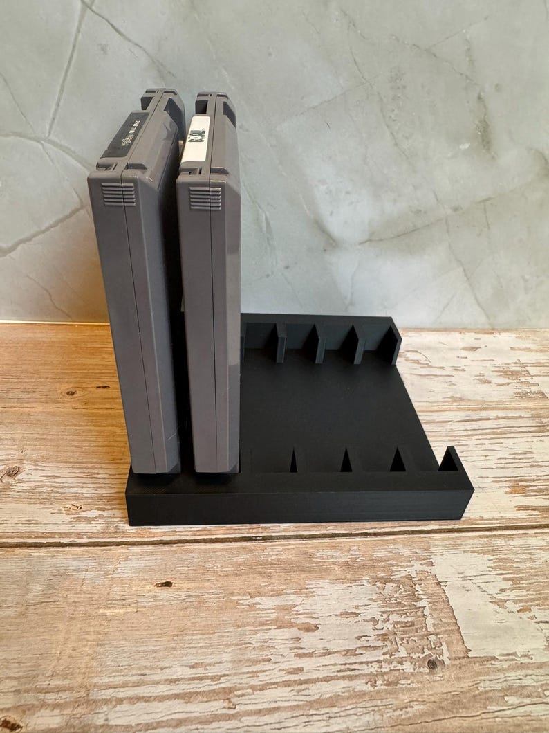 May include: Black, 3D-printed video game cartridge holder. The holder has slots for multiple cartridges and is designed to hold them upright. Two gray game cartridges are in the holder. The holder is on a wooden surface.