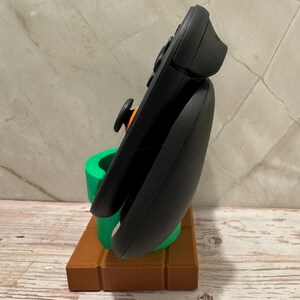 May include: A black games controller is displayed on a 3D-printed stand. The stand features a brown brick base and a green pipe, reminiscent of a classic video game. The controller has an orange joystick and black buttons.