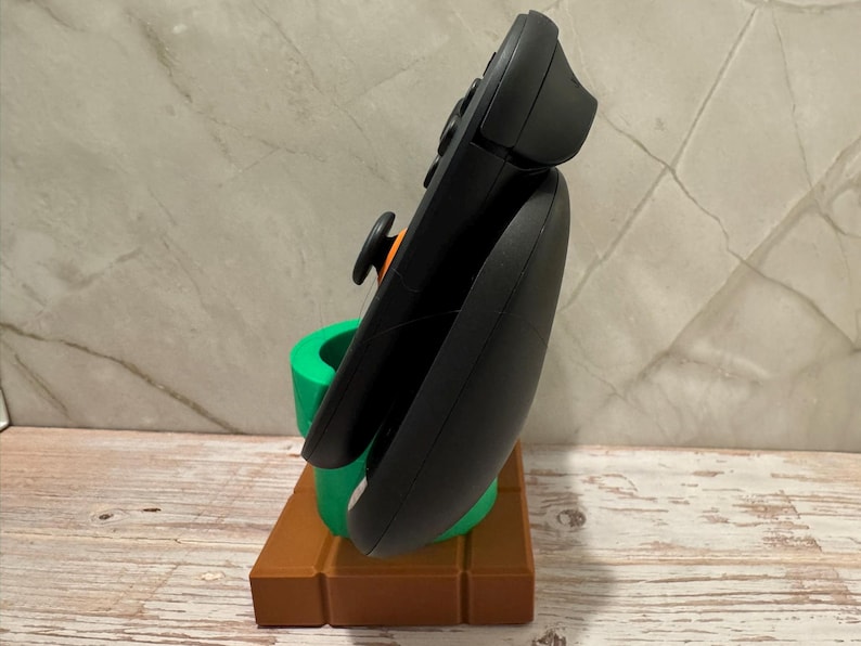 May include: A black games controller is displayed on a 3D-printed stand. The stand features a brown brick base and a green pipe, reminiscent of a classic video game. The controller has an orange joystick and black buttons.