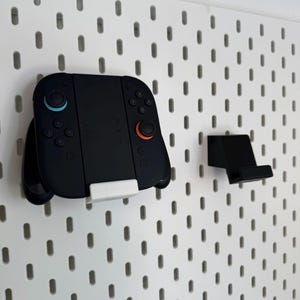 Op de afbeelding: Zwarte gamecontroller met blauwe en oranje knoppen, gemonteerd op een wit pegboard. Een aparte zwarte standaard is ook bevestigd. De controller wordt op zijn plaats gehouden door een witte beugel. Het pegboard heeft gelijkmatig verdeelde ovale gaten.