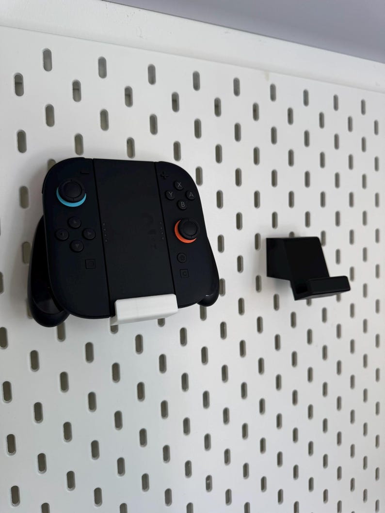 Op de afbeelding: Zwarte gamecontroller met blauwe en oranje knoppen, gemonteerd op een wit pegboard. Een aparte zwarte standaard is ook bevestigd. De controller wordt op zijn plaats gehouden door een witte beugel. Het pegboard heeft gelijkmatig verdeelde ovale gaten.