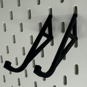 May include: Two black plastic wall mounted hooks with a triangular design. The hooks are mounted on a white pegboard.