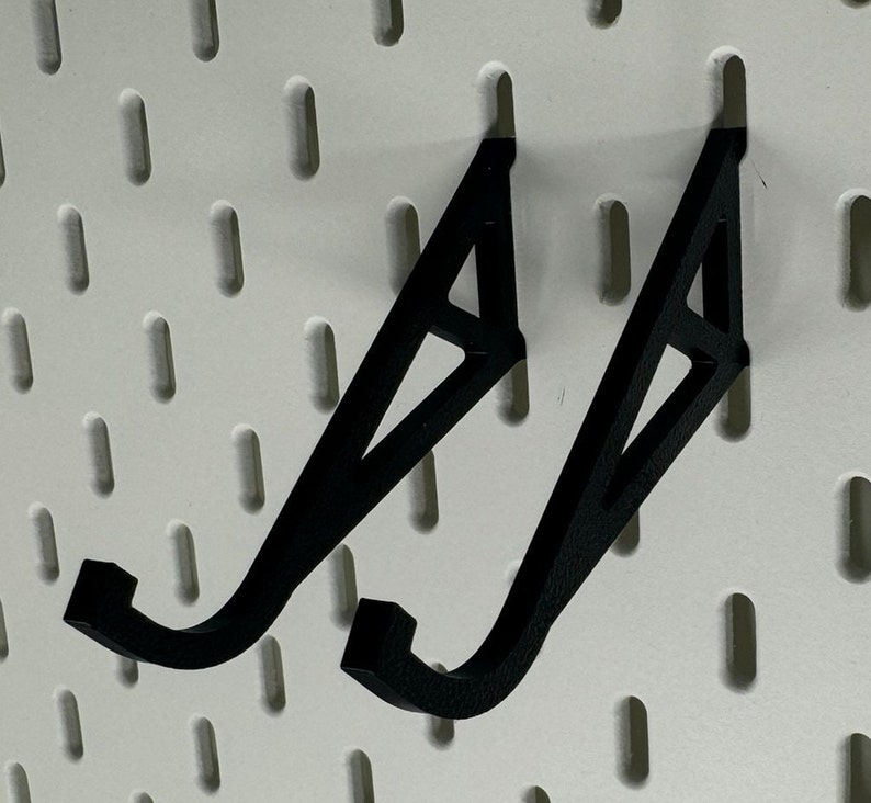May include: Two black plastic wall mounted hooks with a triangular design. The hooks are mounted on a white pegboard.
