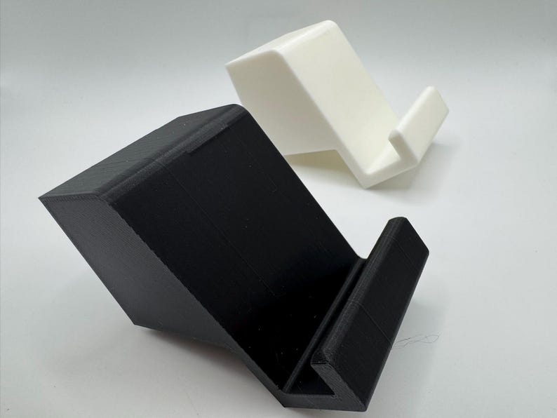 Op de afbeelding: Twee schuine telefoonstandaards, &eacute;&eacute;n zwart en &eacute;&eacute;n wit, worden getoond tegen een witte achtergrond. De standaards hebben een eenvoudig, modern ontwerp met een richel om een telefoon vast te houden en een schuine achterkant ter ondersteuning.