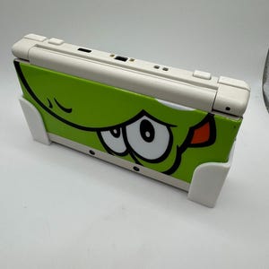 Puede incluir: Consola de juegos port&aacute;til Nintendo 3DS blanca y verde. La cubierta verde presenta la cara de un personaje de dibujos animados con grandes ojos blancos, contornos negros y una lengua roja. La consola est&aacute; soportada por dos soportes blancos.