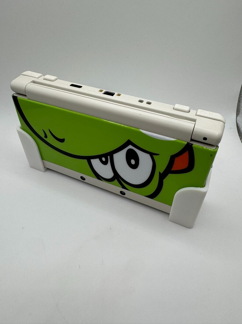 Puede incluir: Consola de juegos port&aacute;til Nintendo 3DS blanca y verde. La cubierta verde presenta la cara de un personaje de dibujos animados con grandes ojos blancos, contornos negros y una lengua roja. La consola est&aacute; soportada por dos soportes blancos.