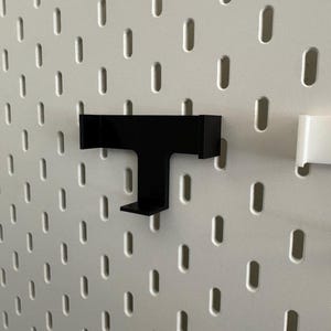 K&ouml;nnte beinhalten: Ein schwarzer, T-f&ouml;rmiger Regalaufsatz f&uuml;r eine Lochwand. Das Regal ist an einer wei&szlig;en Lochwand mit gleichm&auml;&szlig;ig verteilten ovalen L&ouml;chern befestigt. Ein wei&szlig;er Regalaufsatz ist ebenfalls sichtbar.