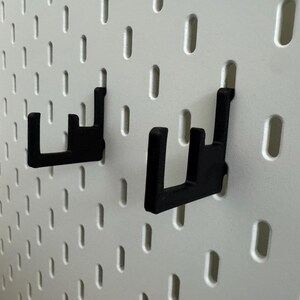 May include: Two black pegboard hooks are mounted on a white pegboard. The hooks are made of a sturdy material and are designed to hold items. The pegboard has evenly spaced holes for versatile storage solutions.