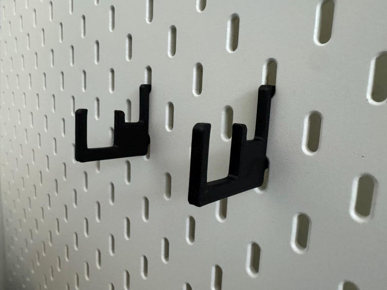 May include: Two black pegboard hooks are mounted on a white pegboard. The hooks are made of a sturdy material and are designed to hold items. The pegboard has evenly spaced holes for versatile storage solutions.
