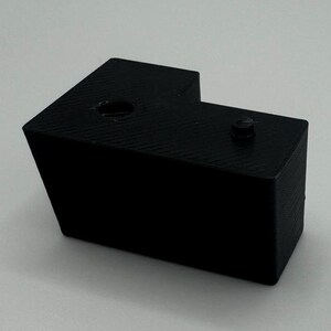May include: A black plastic corner bracket with two holes. The bracket is designed to connect two pieces of wood or other materials at a 90-degree angle.