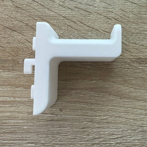 Pu&ograve; includere: Una staffa ad angolo retto in plastica bianca con un'estensione a forma di gancio. La staffa ha una base rettangolare e un braccio perpendicolare con un'estremit&agrave; curva. Presenta due piccole sporgenze rettangolari sul lato.