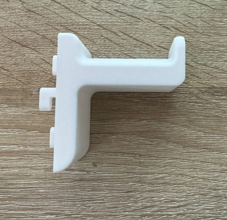 Pu&ograve; includere: Una staffa ad angolo retto in plastica bianca con un'estensione a forma di gancio. La staffa ha una base rettangolare e un braccio perpendicolare con un'estremit&agrave; curva. Presenta due piccole sporgenze rettangolari sul lato.