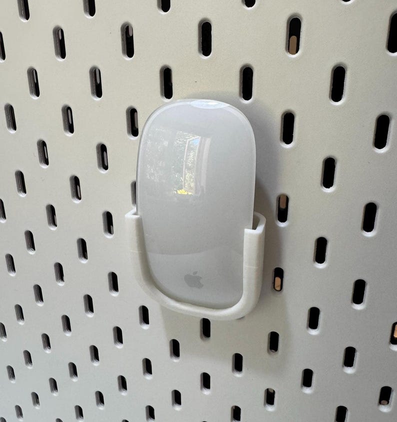 K&ouml;nnte beinhalten: Eine wei&szlig;e Apple Magic Mouse wird in einem wei&szlig;en Kunststoffhalter gehalten, der an einer wei&szlig;en Lochwand befestigt ist. Die Maus hat eine glatte, reflektierende Oberfl&auml;che mit dem Apple-Logo. Die Lochwand hat ein Gitter aus gleichm&auml;&szlig;ig verteilten L&ouml;chern.