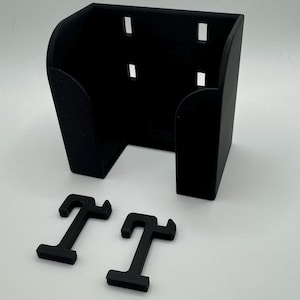 May include: A black, 3D-printed wall mount with a rectangular shape and curved sides. It features several rectangular cutouts and includes two separate, T-shaped mounting brackets. The items are displayed on a white surface.
