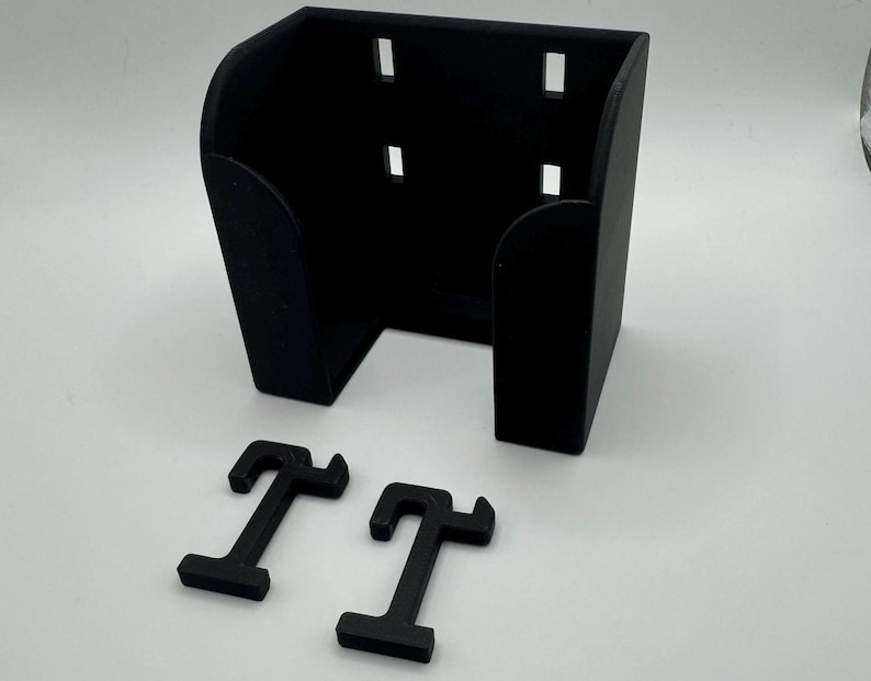 May include: A black, 3D-printed wall mount with a rectangular shape and curved sides. It features several rectangular cutouts and includes two separate, T-shaped mounting brackets. The items are displayed on a white surface.