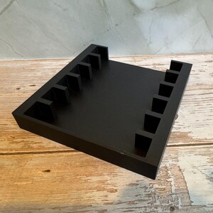 May include: A black wooden organizer with multiple slots, likely for holding items. The organizer is rectangular with a flat base and vertical dividers. The wood grain surface provides a contrast to the black finish.