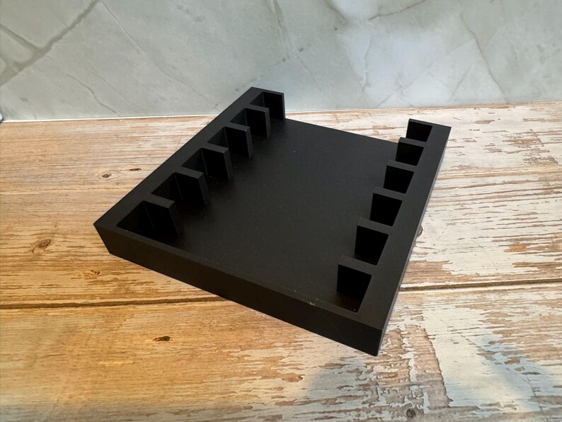 May include: A black wooden organizer with multiple slots, likely for holding items. The organizer is rectangular with a flat base and vertical dividers. The wood grain surface provides a contrast to the black finish.