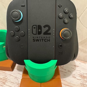 May include: Black Nintendo Switch controller with blue and orange joysticks, and white X, Y, A, and B buttons. The controller is on a green and brown stand, resembling a pipe.