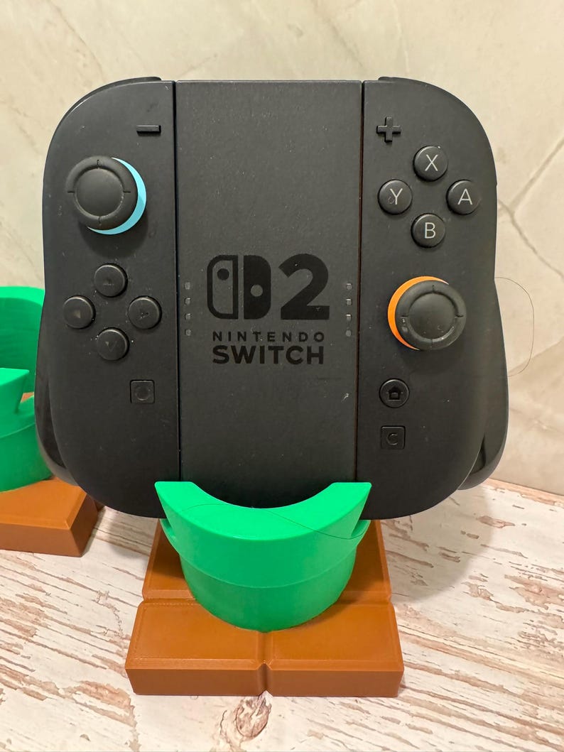 May include: Black Nintendo Switch controller with blue and orange joysticks, and white X, Y, A, and B buttons. The controller is on a green and brown stand, resembling a pipe.