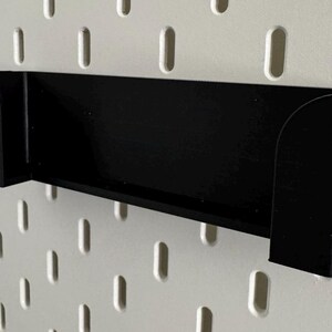 Op de afbeelding: Zwarte plastic wandplank met afgeronde randen. De plank is gemonteerd op een wit prikbord.