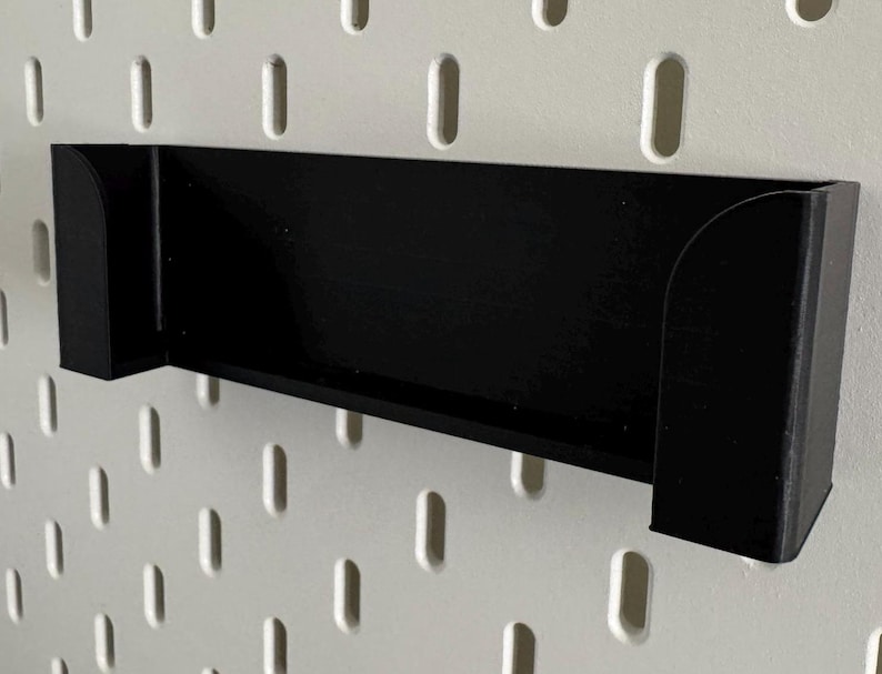 Op de afbeelding: Zwarte plastic wandplank met afgeronde randen. De plank is gemonteerd op een wit prikbord.