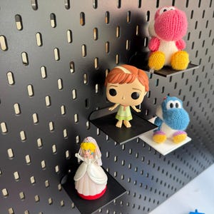 Pode incluir: Um quadro de pinos preto com quatro prateleiras que seguram v&aacute;rias figuras. A prateleira superior tem um Yoshi de malha rosa, a segunda prateleira tem uma figura de menina de cabelo ruivo, a terceira prateleira tem um Yoshi de malha azul e a prateleira inferior tem uma figura de princesa com um vestido branco e cabelo amarelo.