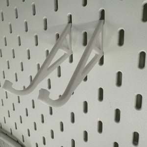 May include: Two white plastic shelf brackets mounted on a white pegboard wall.