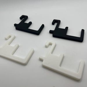 K&ouml;nnte beinhalten: Vier Kunststoffobjekte in Hakenform. Zwei sind schwarz und zwei sind wei&szlig;. Die Objekte haben eine rechteckige Basis mit einem nach oben verlaufenden Haken und einer rechtwinkligen Verl&auml;ngerung. Die Artikel dienen wahrscheinlich zum Aufh&auml;ngen oder Sichern.