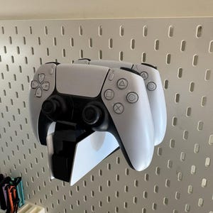 K&ouml;nnte beinhalten: Zwei wei&szlig;e und schwarze Gamecontroller sind an einer wei&szlig;en Lochwand befestigt. Die Controller sind &uuml;bereinander gestapelt, wobei die Kn&ouml;pfe und Joysticks sichtbar sind. Die Lochwand hat ein Raster aus L&ouml;chern.